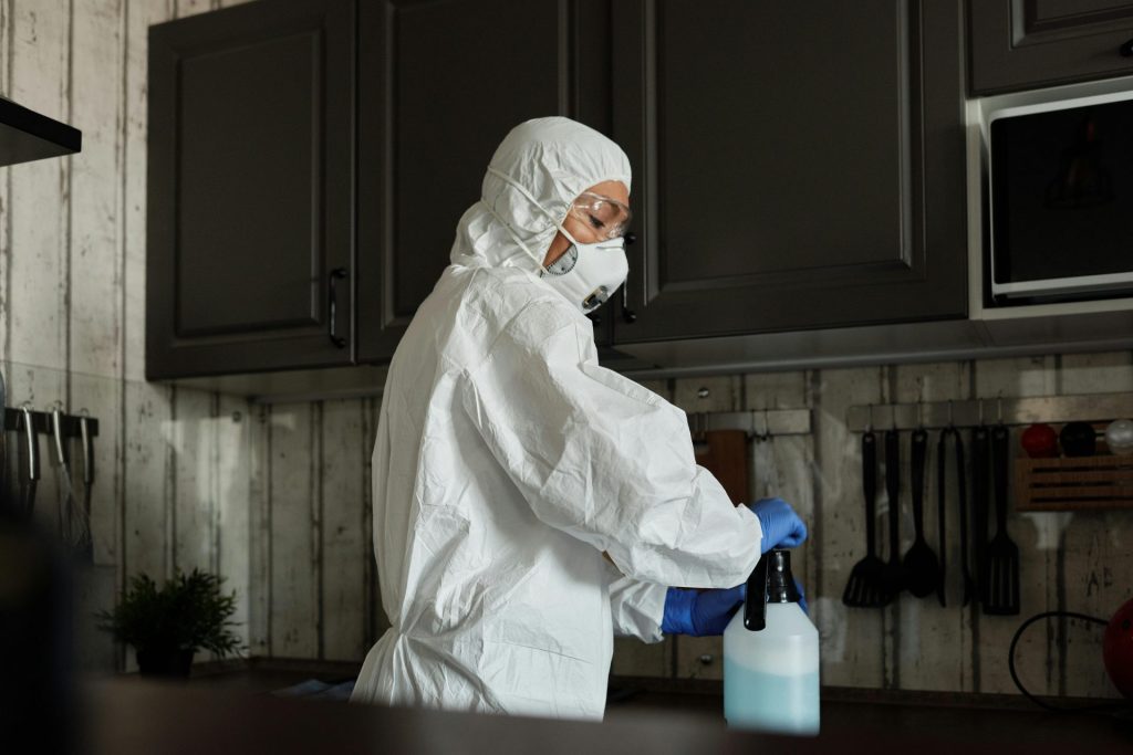Ein Mitarbeiter des Gesundheitswesens in persönlicher Schutzausrüstung verwendet während der COVID-19-Pandemie in einer Küche Desinfektionsspray.