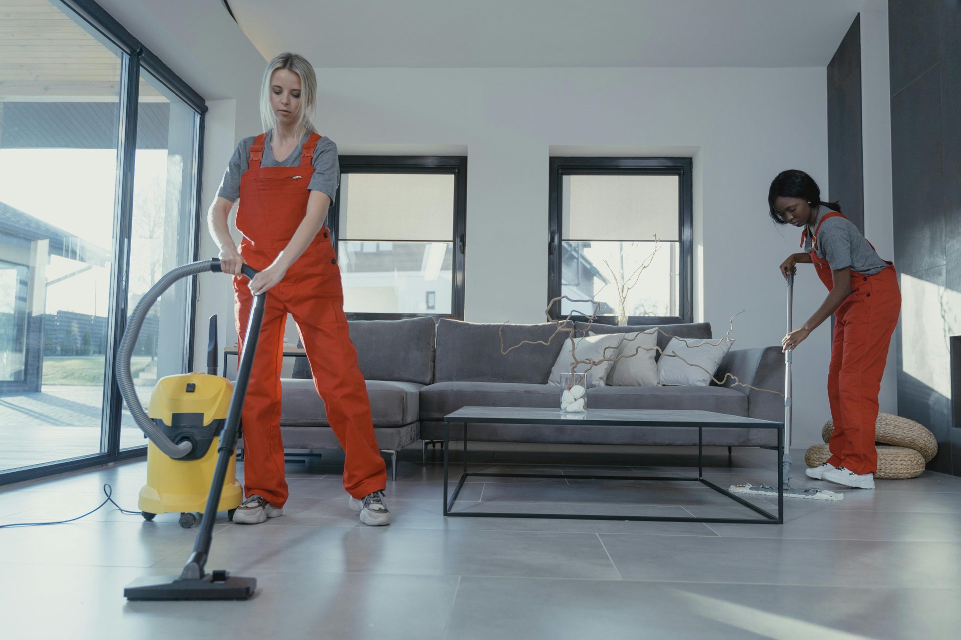 Zwei Frauen in roten Overalls saugen und fegen ein modernes Wohnzimmer.