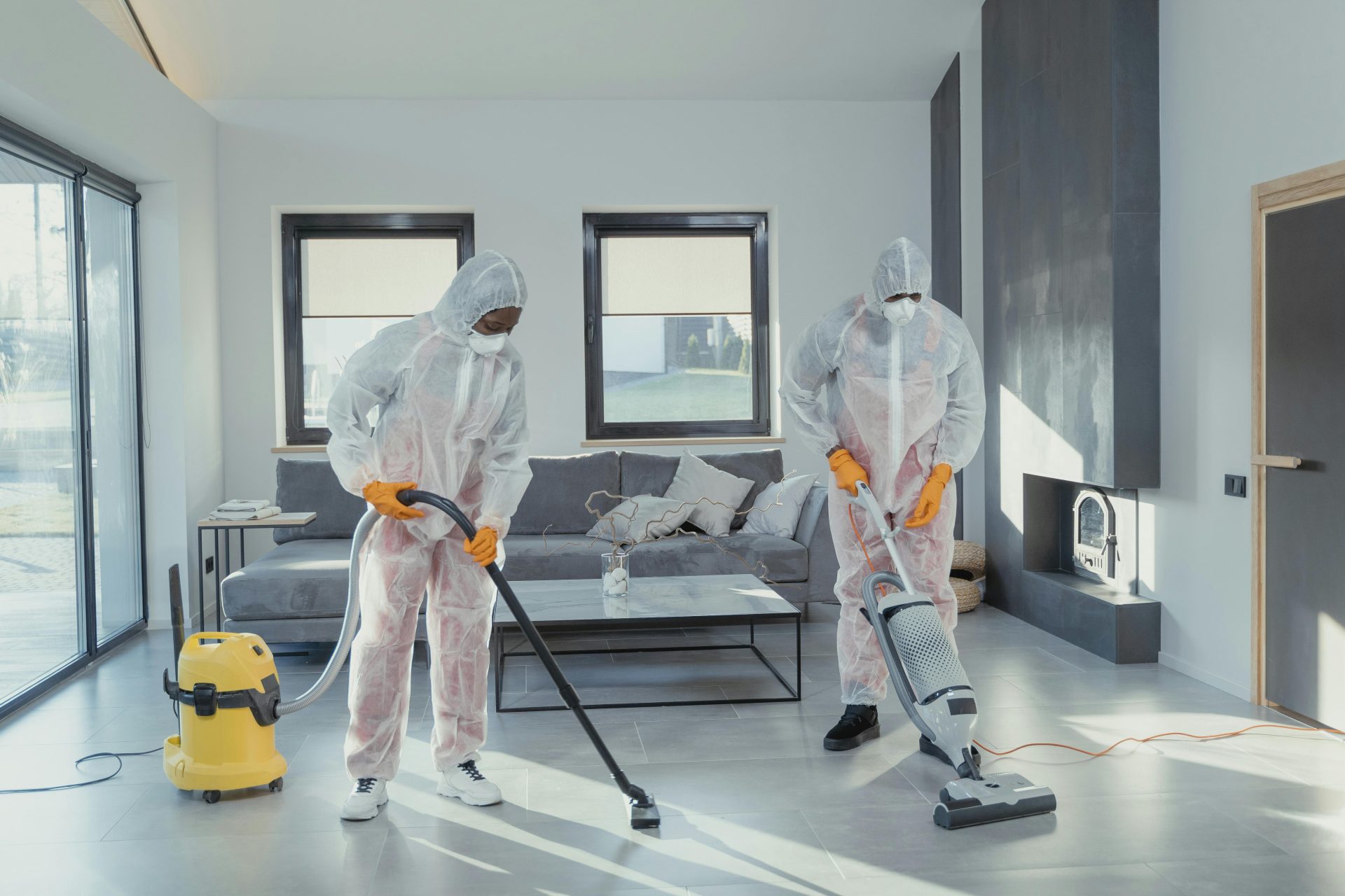 Zwei Arbeiter in Schutzkleidung saugen und desinfizieren ein modernes Wohnzimmer.