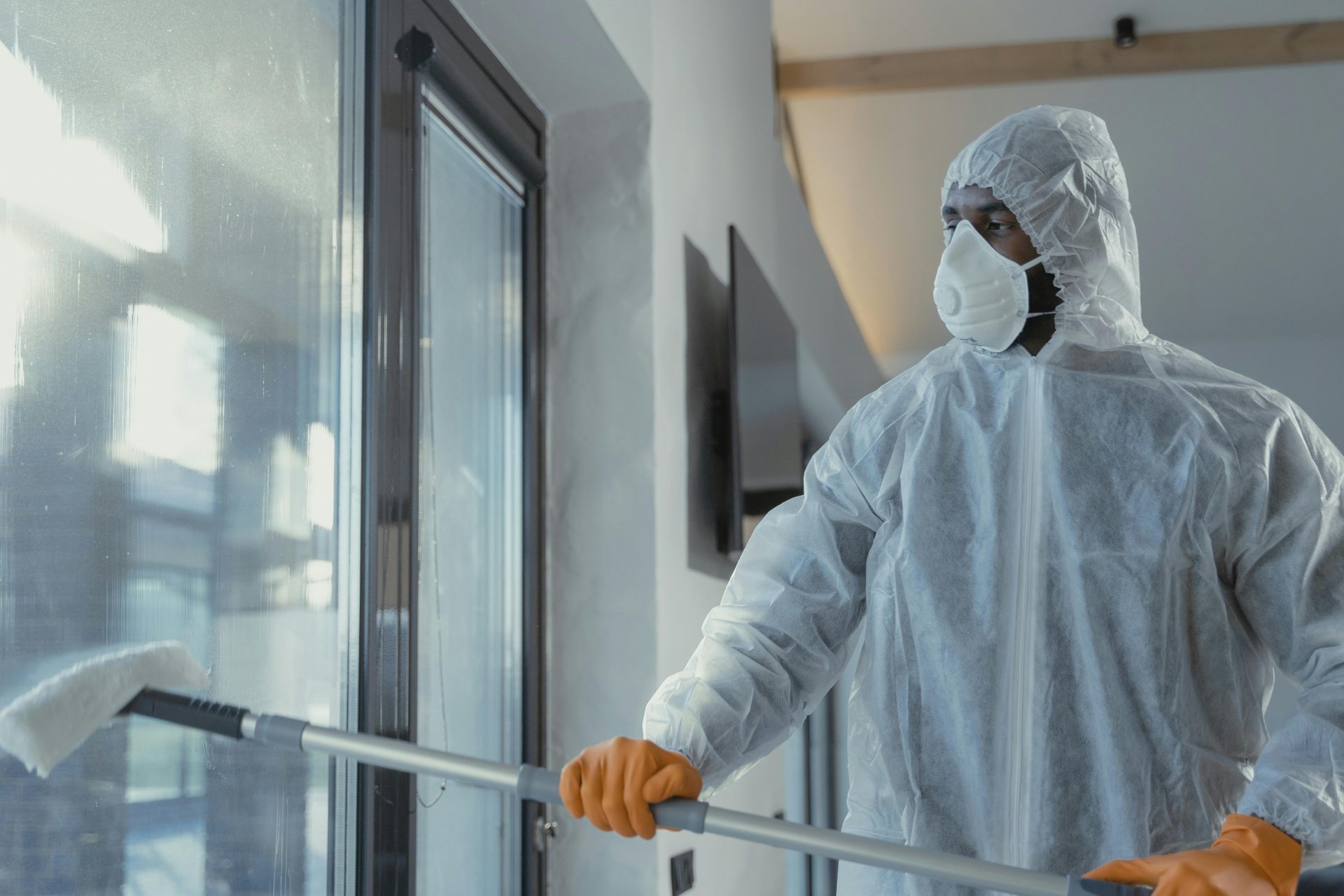 Erwachsener männlicher Reinigungskraft in Schutzanzug und Maske putzt an einem sonnigen Tag drinnen Fenster.