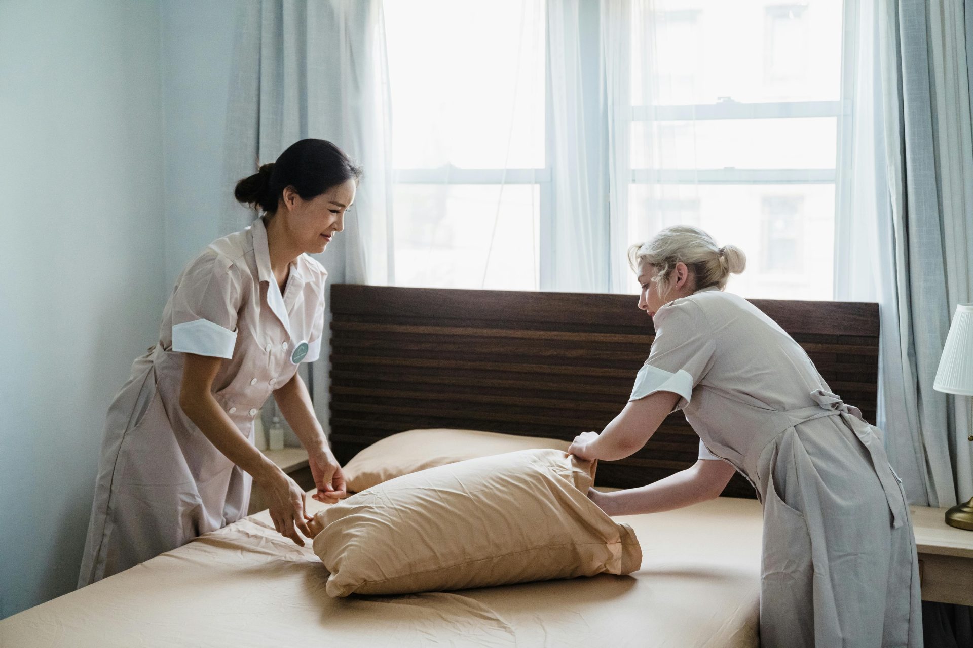 Zwei Haushälterinnen in Uniform arbeiten zusammen, während sie in einem hellen, ruhigen Schlafzimmer ein Bett machen.